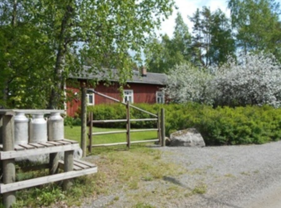 Ehtoohartaus Helinin torpalla Helinin torppa eli punainen vanha puutalo, jonka ympärillä vehreitä puita ja pensaita, etualalla vanhat maitotonkat maitolaiturilla.